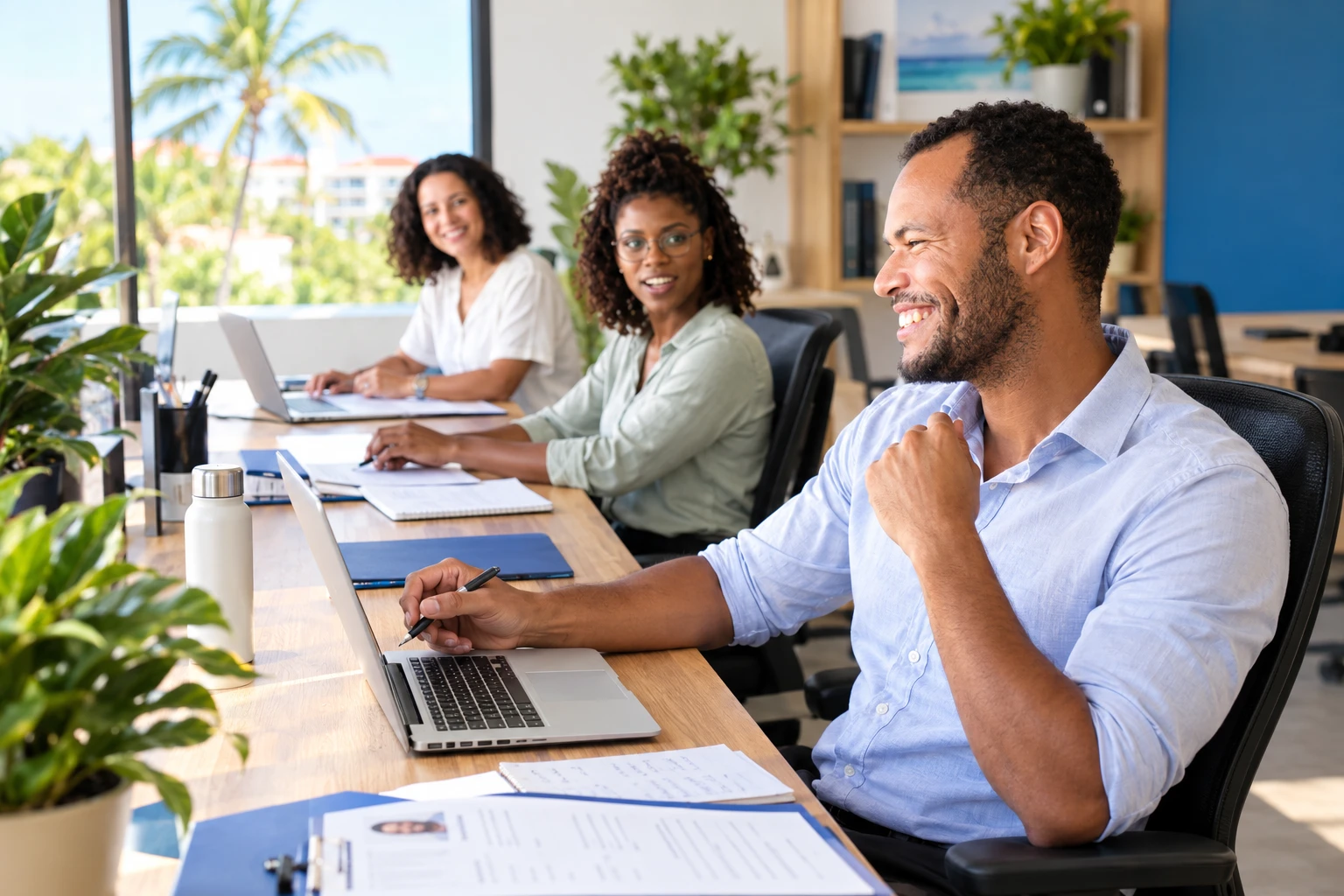 Three colleagues smiling at their desks in a bright office with palm trees outside the window